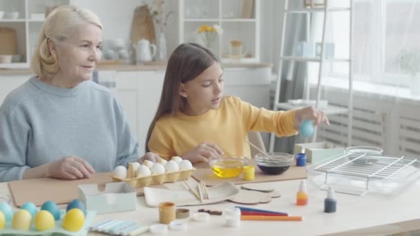 青い食品着色料とボウルから染色卵を取り キッチンテーブルで祖母と一緒にイースターの装飾を作りながら 乾燥棚に置きます — ストック動画