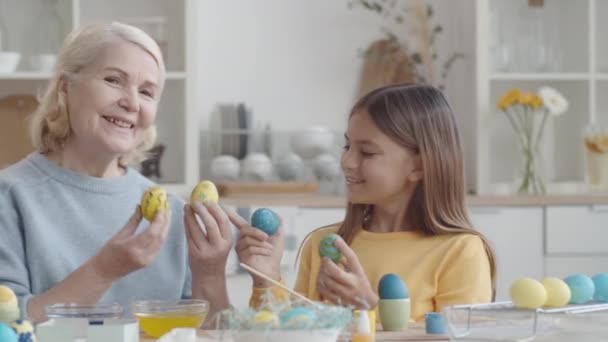 喜ばしい祖母と小さな孫娘の撮影を追跡染色イースターエッグと笑顔でカメラを見ながらキッチンテーブルに座って — ストック動画