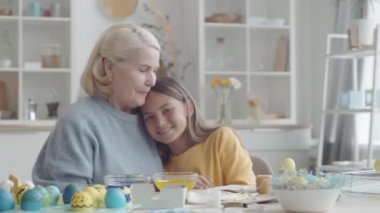 Sevimli küçük bir kız ve mutlu bir büyükanne birbirlerine sarılıyor, gülümsüyor ve üzerinde paskalya yumurtası olan mutfak masasında birlikte otururken kameraya bakıyor.