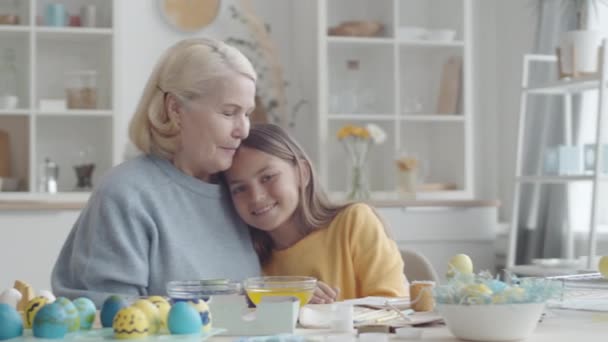 素敵な女の子と幸せなシニア祖母が抱き合って 笑顔でカメラを見て イースターエッグのキッチンテーブルに座っています — ストック動画