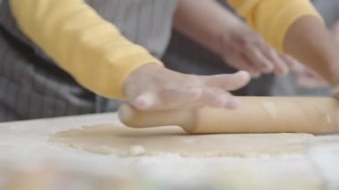 Mutfak masasında büyükannesiyle yemek pişirirken hamur işlerini ahşap broşla yapan önlüklü küçük kızın ellerini yakın çekimde çek.