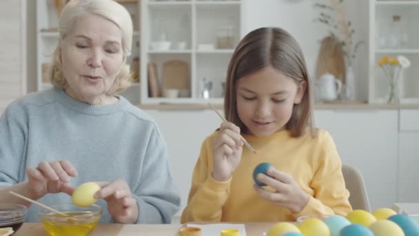 台所のテーブルに座っている陽気な先輩の女性は 食べ物の着色で卵を浸し 彼女はブラシで卵を染色上のパターンを描画しながら 小さな孫娘と話 — ストック動画