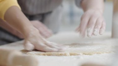 Anneannenin ve küçük torununun hamur işlerinde un ellerken ve masanın üzerinde ahşap bir iğne ile yuvarlarken yakın plan çekimleri.
