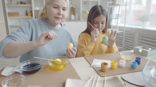 放大照片拍摄的11岁女孩和老奶奶在厨房用画笔和水彩画彩绘复活节彩蛋时 笑着和聊天 — 图库视频影像