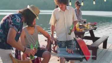 Izgaranın yanında duran, tost şişelerini tokuşturan ve yaz günü göl partisi yaparken bira içen çok ırklı erkek arkadaş topluluğu.