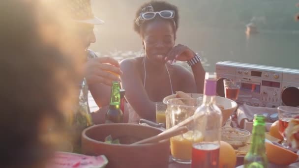 友人と夕食のテーブルに座っている若い中東の男性とアフリカ系アメリカ人の女性 夏の日に湖のパーティー中に笑顔とチャット — ストック動画