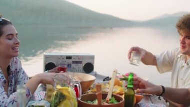 Yaz günü göl partisinde iskelede düzenlenen şenlikli yemekte kadehleri tokuşturup kadeh tokuşturan çok ırklı genç insanların görüntüsünü büyüt.