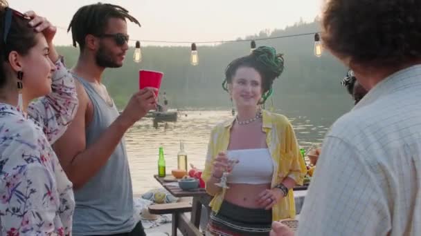 夏の夜に湖のパーティーをしながら ガーランドで飾られた桟橋で円の中でおしゃべり 飲み物やダンスの若い幸せな多民族の男性と女性のパンニングショット — ストック動画
