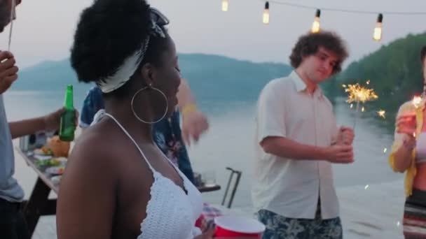 興奮したアフリカ系アメリカ人の女性は 夕方に湖のパーティーをしながら 多民族の友人の会社とカップ 笑顔とダンスを開催 — ストック動画