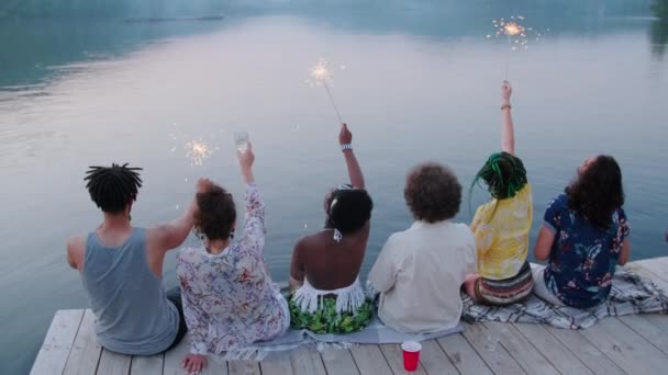 夕阳西下时 年轻的多民族朋友坐在木制码头边 手握火花 挥动着胳膊参加湖畔派对 倒立在后视镜下的镜头 — 图库视频影像
