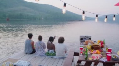 Göl manzarasından hoşlanan, içki içen ve sohbet eden, ahşap iskelede, üzerinde yemek masası ve çelenk olan bir arada oturan genç erkek ve kadınların arka plandaki görüntüsü.