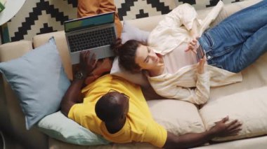 Kanepede oturup dizüstü bilgisayarda sörf yapan Afro-Amerikan bir adamın karısı ona yaslanırken ve akıllı telefon kullanırken en iyi görüntüsü.