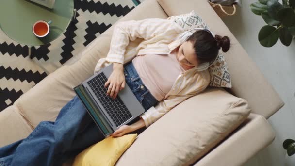 在家里躺在沙发上用无线耳机在笔记本电脑上打字的黑发女人的自上而下的照片 — 图库视频影像