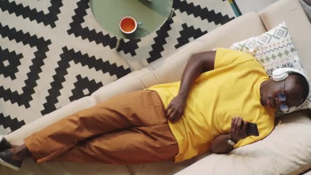 年轻的非洲裔美国人头戴无线耳机 躺在沙发上 一边用智能手机听音乐 一边带着节奏地摇头 高透视 — 图库视频影像
