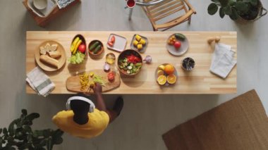 Kablosuz kulaklık takan, salata için sebze kesen ve evde yemek pişirirken dans eden Afro-Amerikalı adamın yukarıdan görünüşü