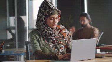 Başörtülü güzel Müslüman iş kadını portresi ofis masasında dizüstü bilgisayarda çalışıyor ve kameraya özgüvenle bakıyor.