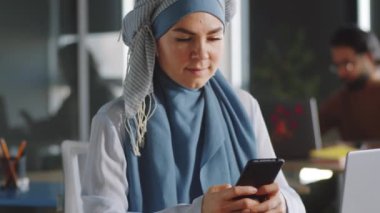 Portre, genç, pozitif Müslüman iş kadınının tesettürlü akıllı telefondan mesajlaşmasını ve iş günü kameraya poz vermesini gösteriyor.