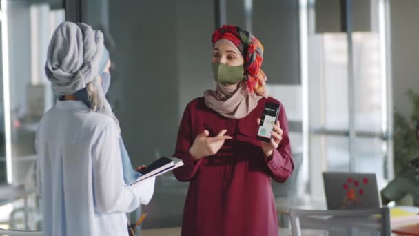 ヒジャーブ州のイスラム教徒のビジネス女性とオフィスに立ってマスクに直面し コロナウイルス発生時に働く女性同僚とスマートフォンのプレゼンテーションを議論 — ストック動画