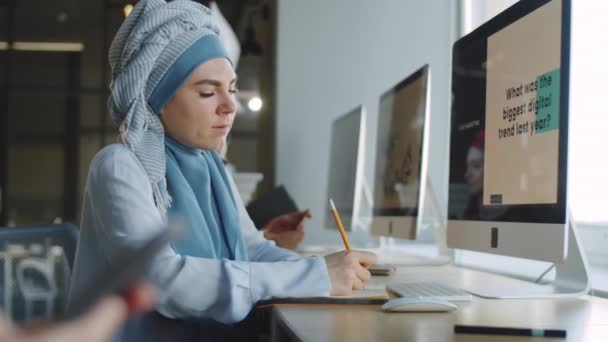 ヒジャーブの若いイスラム教徒女性のサイドビューコンピュータ上のビジネスプレゼンテーションを見て オフィスで働いている間にメモを取る — ストック動画