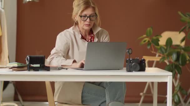 在工作室里 穿着时髦休闲装 戴着眼镜 在笔记本电脑上工作的时髦老年女性的倒立照片 — 图库视频影像