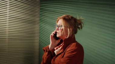 Göğüs hizasında şık giyinmiş güzel bir iş kadınının cep telefonuyla konuşurken pencerenin yanında panjurlarla dikilirkenki görüntüsü.