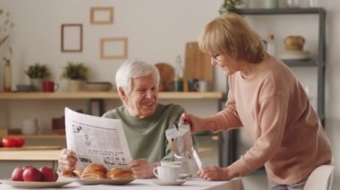 Gazete tutan ve karısıyla konuşan kıdemli bir adam kahve fincanlarına kahve dökerken ve mutfak masasında onun yanında otururken