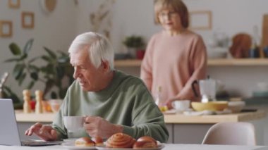 Üst düzey beyaz adamın mutfak masasında oturup dizüstü bilgisayarını karıştırırken ve karısı arka planda yemek pişirirken elinde çay fincanı tutarken çekilen görüntüleri.