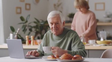 Mutfak masasında dizüstü bilgisayarı ve çay bardağıyla oturan ve kameraya poz veren üst düzey bir adamın portresi.