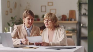 Kıdemli kadın ve kadın finans danışmanı faturaları tartışıyor, sonra birlikte kameraya poz veriyor ve evde buluşurken gülümsüyor.
