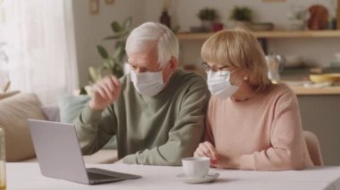 Koruyucu maskeli yaşlı bir çift masada oturuyor, bilgisayar başında video görüşmeleri yapıyor ve Coronavirus salgını sırasında evde otururken kucaklaşıyorlar.