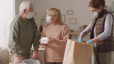 Koruyucu maskeli ve eldivenli bir kadın evde yaşlı bir çift için alışveriş poşetinden market ürünleri indirirken aynı zamanda salgın sırasında gıda alışverişinde olan emeklilere yardım ediyor.