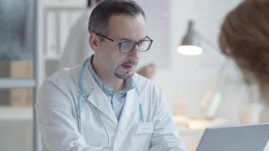 Laboratuar önlüklü, boynunda steteskop olan, dizüstü bilgisayar kullanan ve muayenehanedeki kadın hastaya danışmanlık yapan erkek doktorun omzundan.
