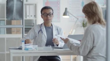 Laboratuvar önlüklü ve gözlüklü melez bir kadın doktor kadınla konuşuyor ve klinikte danışmanlık yaparken imzalaması için ona tıbbi hizmet sözleşmesi veriyor.