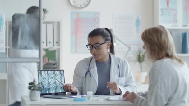 ラップトップ画面に脳のX線画像を表示し 診療所で医療相談中に患者からの質問に答える若い美しい混合レース女性医師 — ストック動画