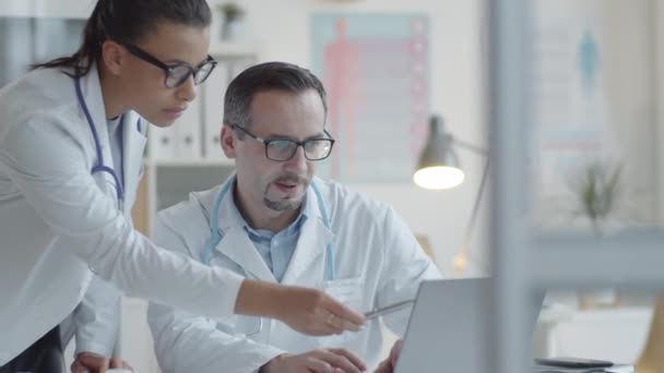 若い混合レース女性医師と白人男性医師は 医療事務所で一緒に働くノートパソコンの画面上で何かを議論 — ストック動画