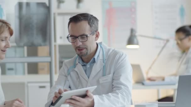 男性医師はデジタルタブレットで何かを示し クリニックでの医療相談中に中年女性患者と話す — ストック動画