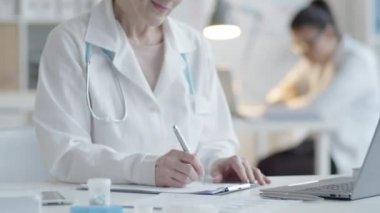 Laboratuvar önlüklü orta yaşlı kadın doktorun çalışma masasında reçete yazarken, kameraya bakıp, iş saatlerinde poz verirken gülümsemesini çek.