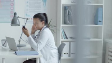 Laboratuvar önlüklü genç melez kadın doktorun yan görüntüsü. Üzerinde dizüstü bilgisayarla masada oturuyor ve sağlık ofisinde çalışırken akıllı telefon kullanıyor.