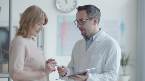 白人男性開業医のウエストアップ女性患者に歩いて 話をし 現代のクリニックで働いている間に署名するための彼女の医療契約を与える — ストック動画