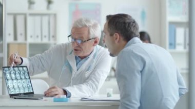 Kıdemli doktor dizüstü bilgisayardaki beyin röntgenine işaret ediyor ve klinikte tıbbi danışmanlık yaparken genç erkek hastaya teşhisi açıklıyor.