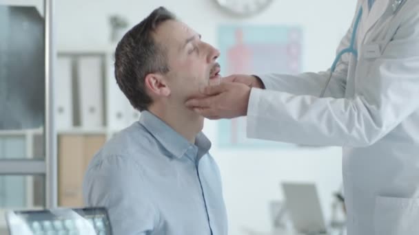 男性患者の首にリンパ節を検査し 診療所での健康診断時に彼に診断を伝えるラップコートで上級医師のショットを傾けます — ストック動画