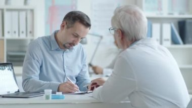 Genç beyaz adam sağlık hizmeti anlaşması imzalıyor ve tıp bürosunu ziyaret ederken kıdemli doktorla tokalaşıyor.