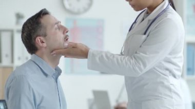 Melez ceketli, erkek hastanın boynundaki lenf düğümlerini inceleyen ve klinikteki sağlık kontrolü sırasında her şeyin yolunda olduğunu söyleyen genç, melez kadın doktoru yukarı kaldır.
