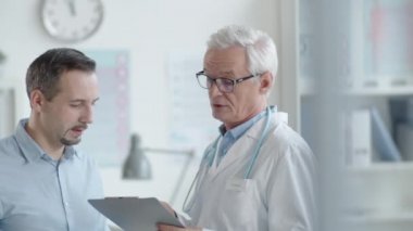 Laboratuvar önlüklü ve gözlüklü kıdemli doktor elinde not panosuyla tıp bürosunda danışmanlık yaparken genç bir erkek hastayla konuşuyor.