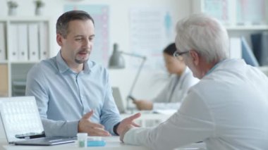 Kafkasyalı genç bir adam muayene masasında oturuyor ve danışma sırasında notlar alırken kıdemli doktora bir şeyler anlatıyor.