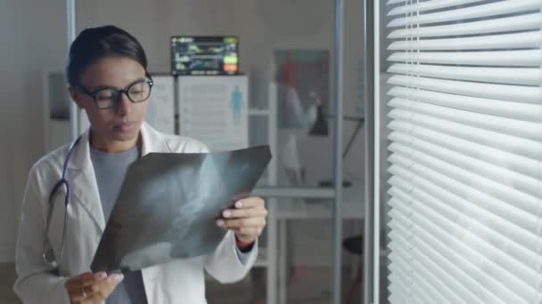 身穿实验室外套 戴着眼镜 头戴百叶窗的年轻混血女医生在医务室检查X光照片 — 图库视频影像
