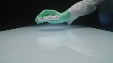 Steril eldivenli ve üniformalı eczacılık laboratuarı çalışanının eline zum yap. Karanlık stüdyodaki beyaz masanın üzerine çeşitli ilaç kapsülleri ve tabletlerle cam kap koy.