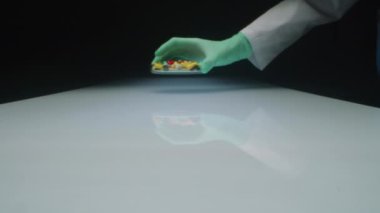 Steril eldivenli ve laboratuvar önlüklü tanımlanamayan sağlık görevlisinin, çeşitli ilaç kapsülleri ve haplarla dolu cam kapları beyaz masanın üzerine yerleştirdiği stüdyoyu yakınlaştır.