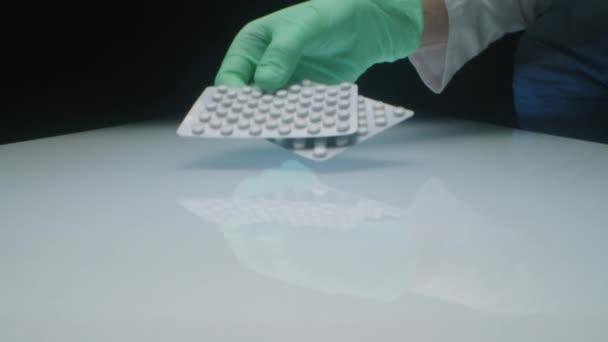 Přiblížit záběr ruky nerozpoznatelného laboratorního pracovníka v jednorázových rukavicích a bílém plášti, který dává dva blistry s tabletami léku na skleněný stůl v tmavém ateliéru