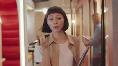 Gündelik kıyafet ve gözlüklü genç bir kadın çalışma odasının koridorunda yürüyor, hoparlörden konuşuyor ve elinde modern bir dizüstü bilgisayar tutuyor.